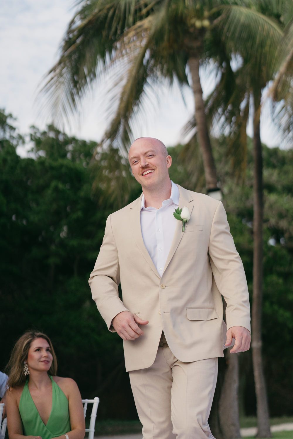 Smiling groom in beige suit with boutonniere walks down outdoor wedding aisle flanked by palm trees.