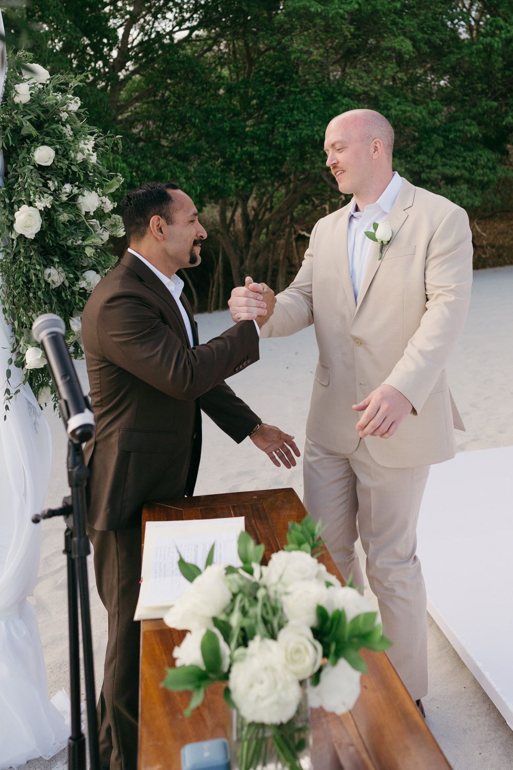 Groom in tan suit shakes hands with officiant at outdoor beach wedding ceremony altar with white flowers.