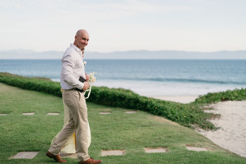 Bald groom in linen suit carries white boutonniere while walking on green lawn with ocean backdrop.