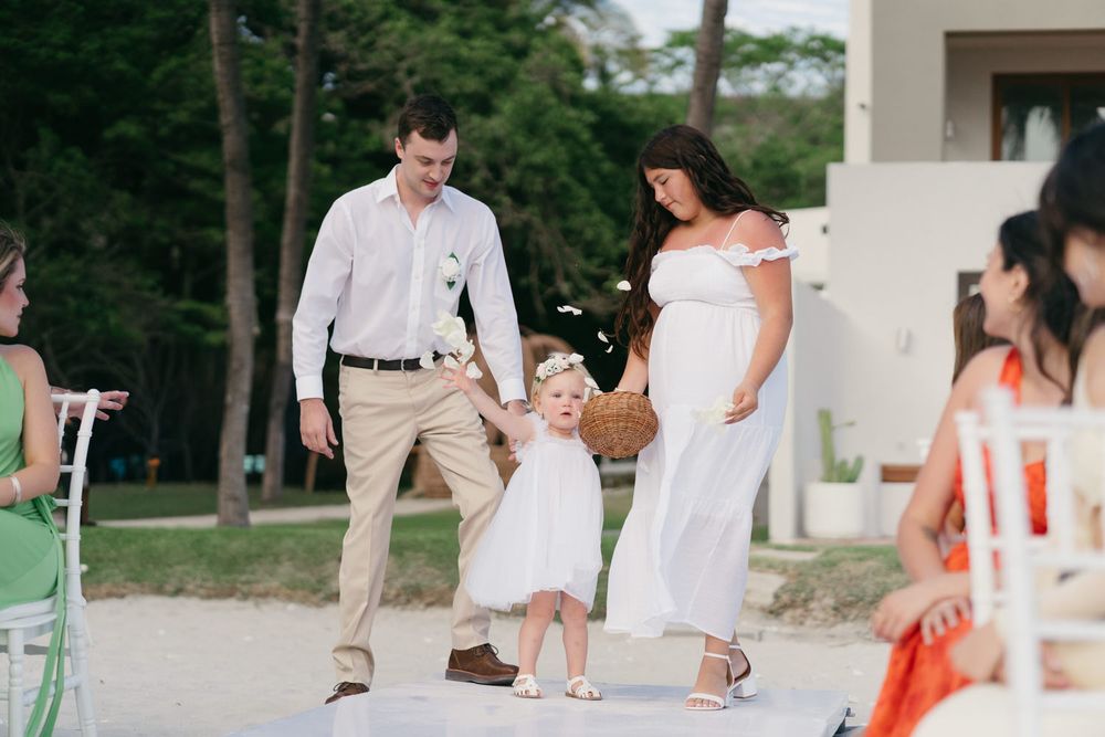 Pregnant woman in white dress walks with husband and flower girl at outdoor wedding ceremony.