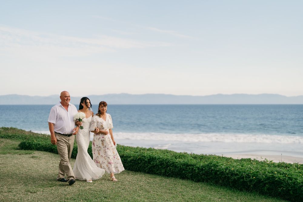 Bride in white gown walks with two guests along oceanside lawn toward beach wedding ceremony.