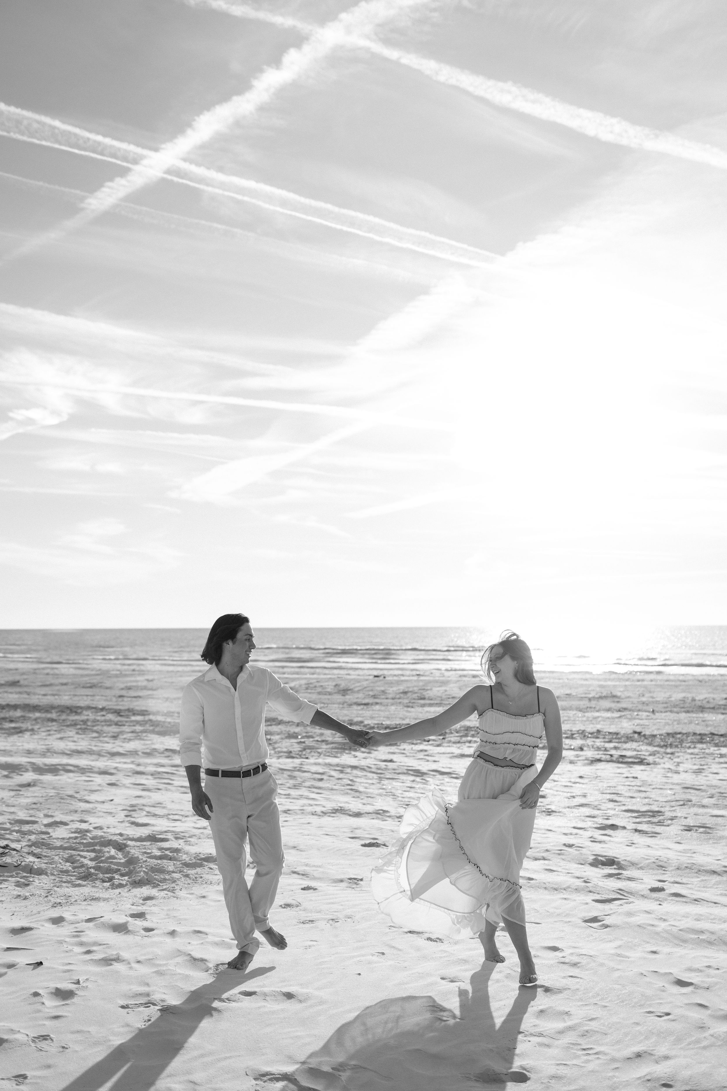 Black and white photo of a couple holding hands and walking on a sunny beach with ocean waves and contrails in the sky.