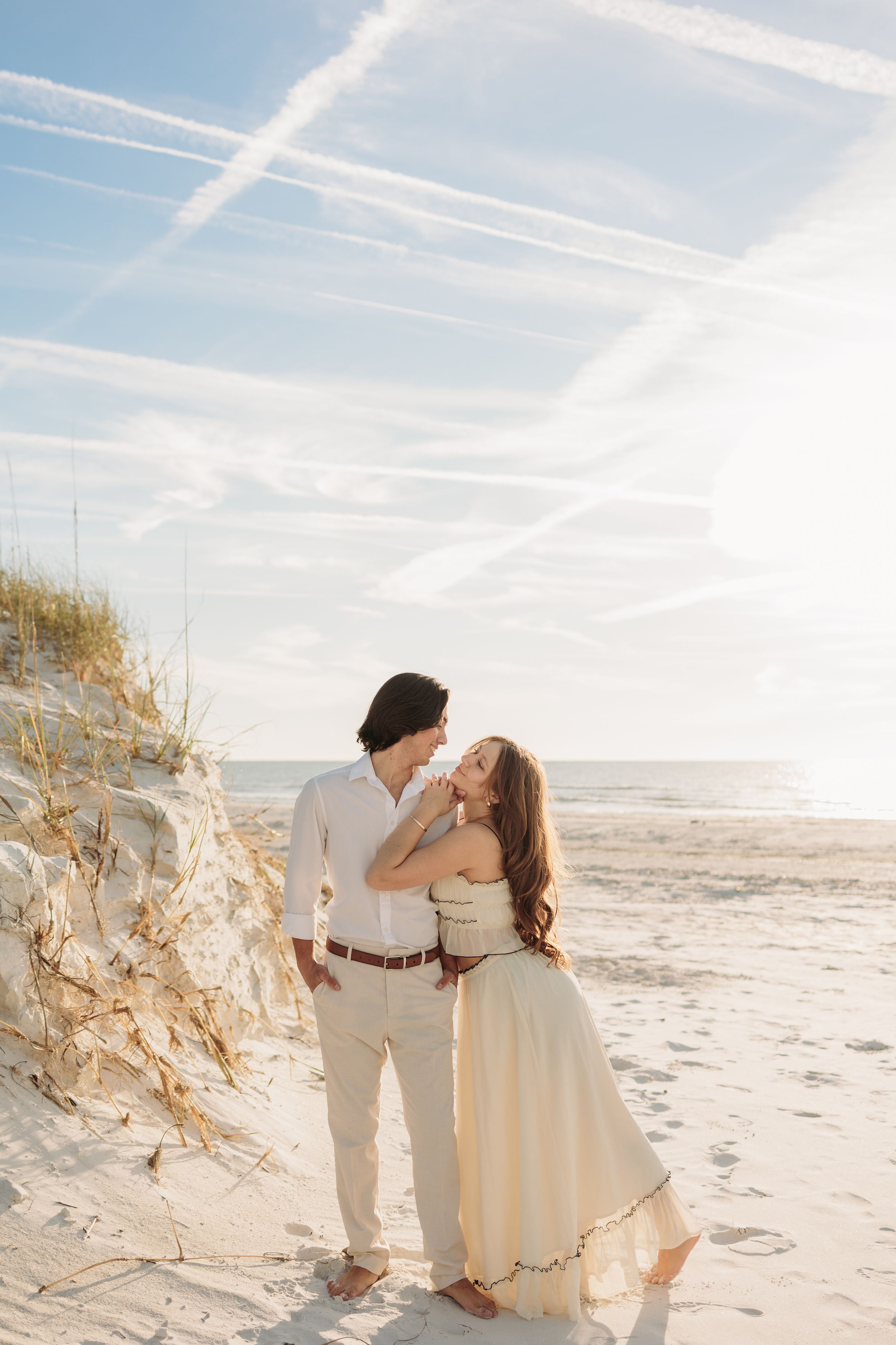 Romantic couple embracing on a sunny beach with sand dunes, wearing cream outfits, with contrails crossing the blue sky.