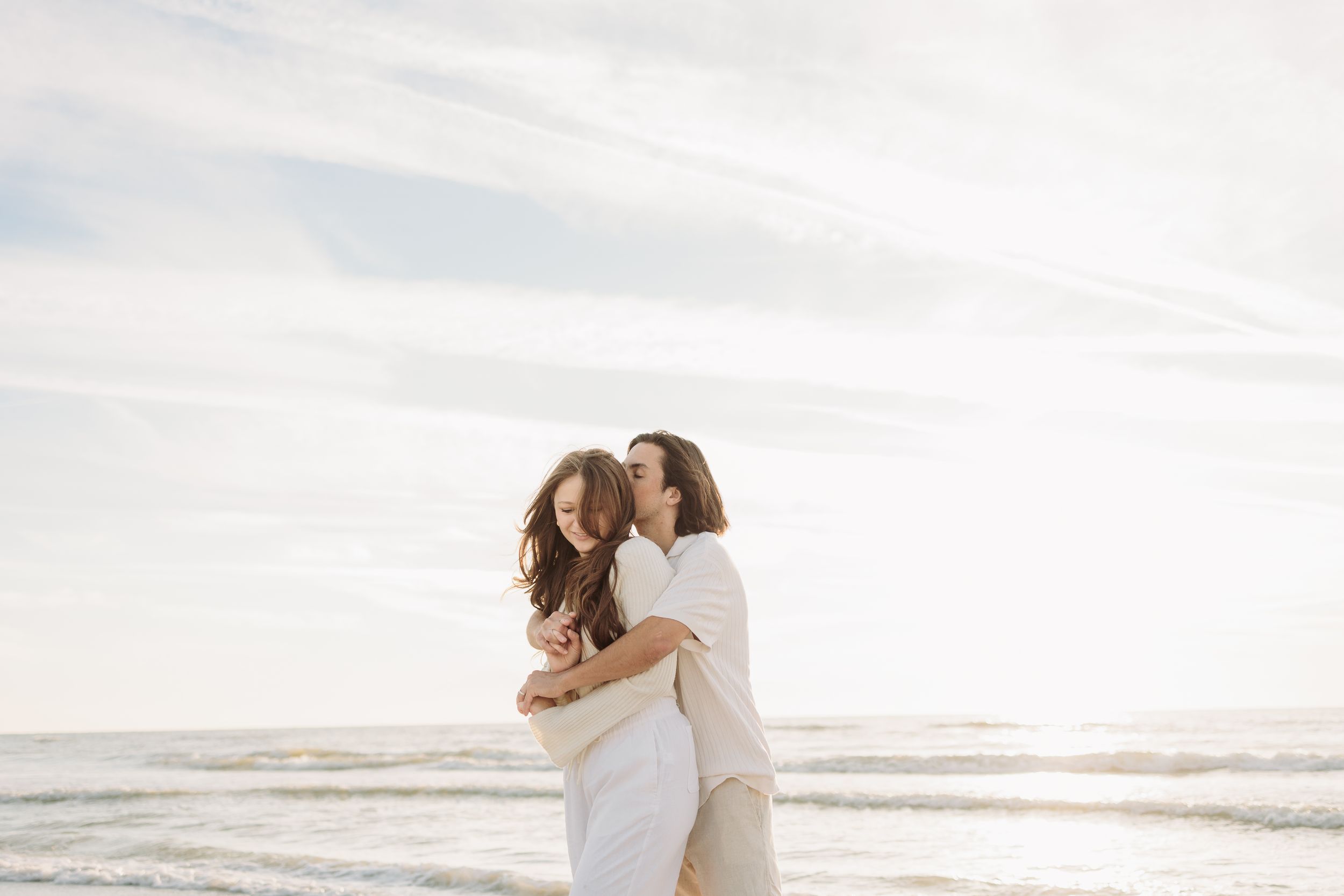 Two women embracing and laughing together on a sunny beach with ocean waves in the background.