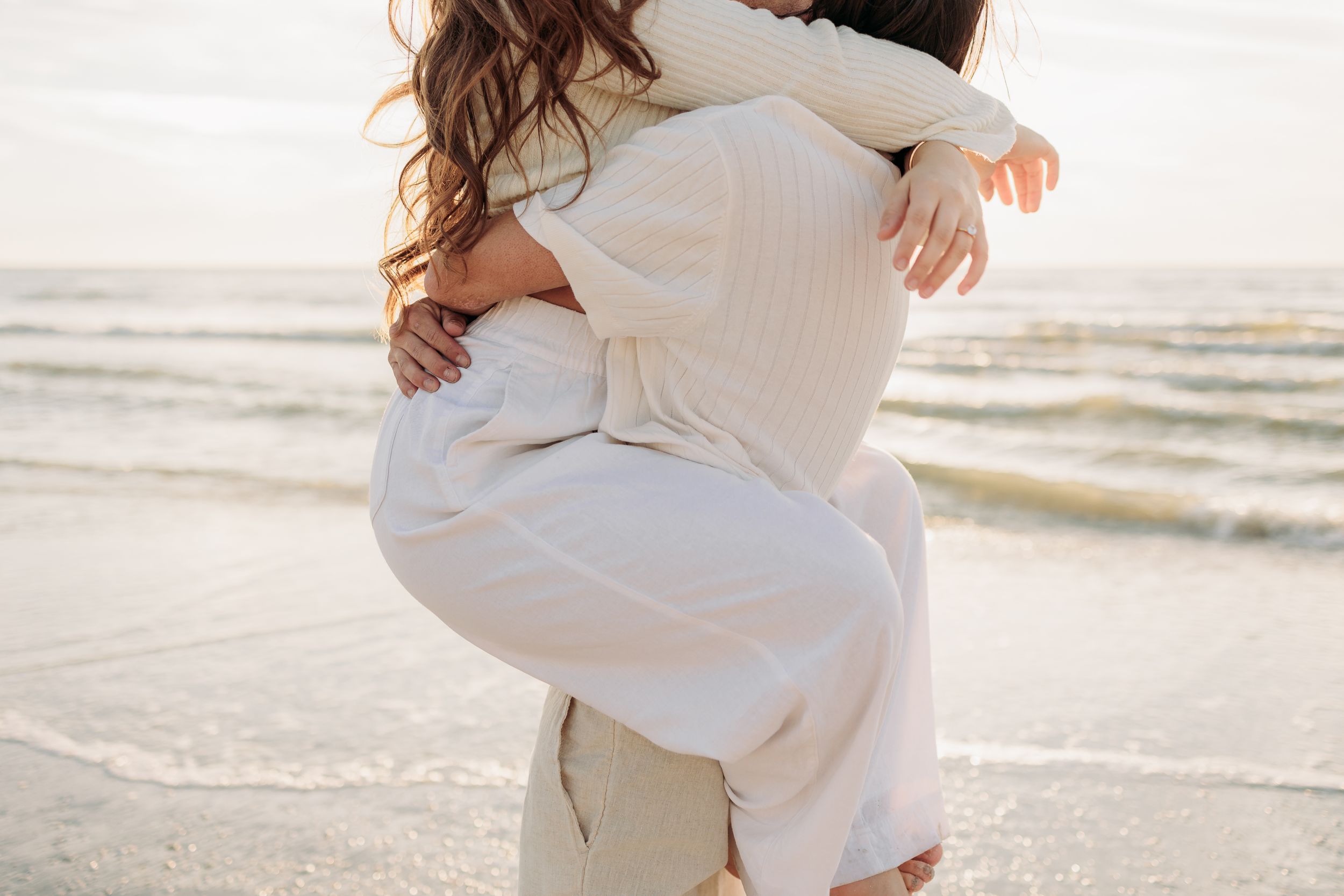 Couple embracing in a romantic hug on a sandy beach at sunset, both wearing white outfits.