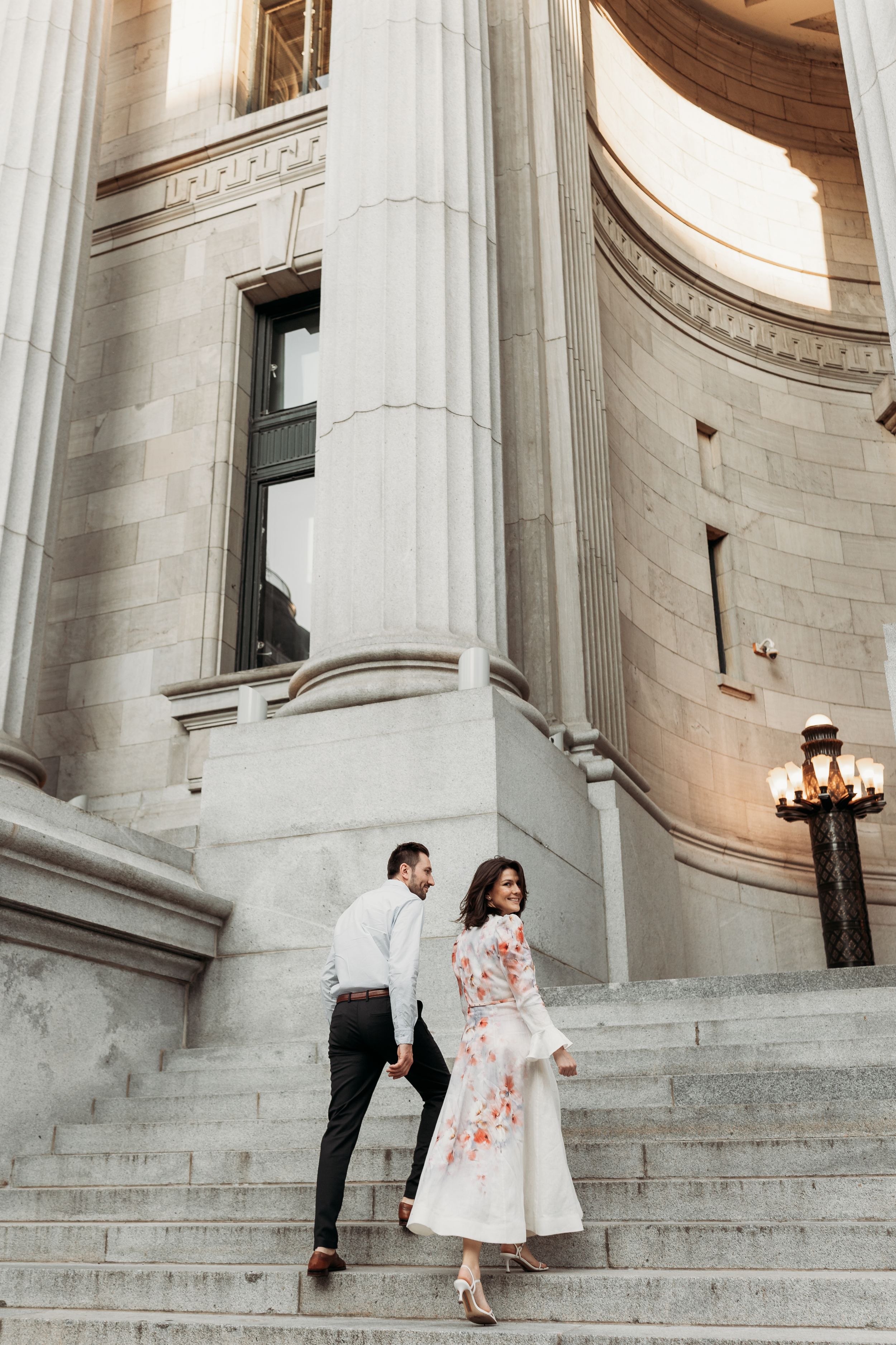 Couple in elegant attire walking up grand stone steps with tall columns at a classical building.
