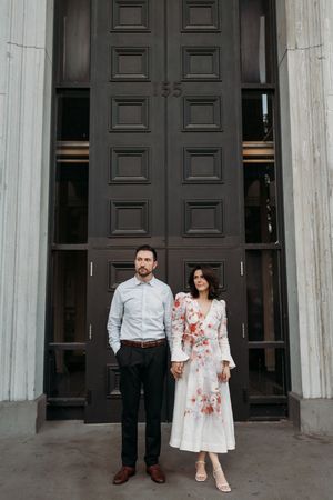 Couple standing hand in hand before large dark wooden doors, man in blue shirt, woman in floral white dress.