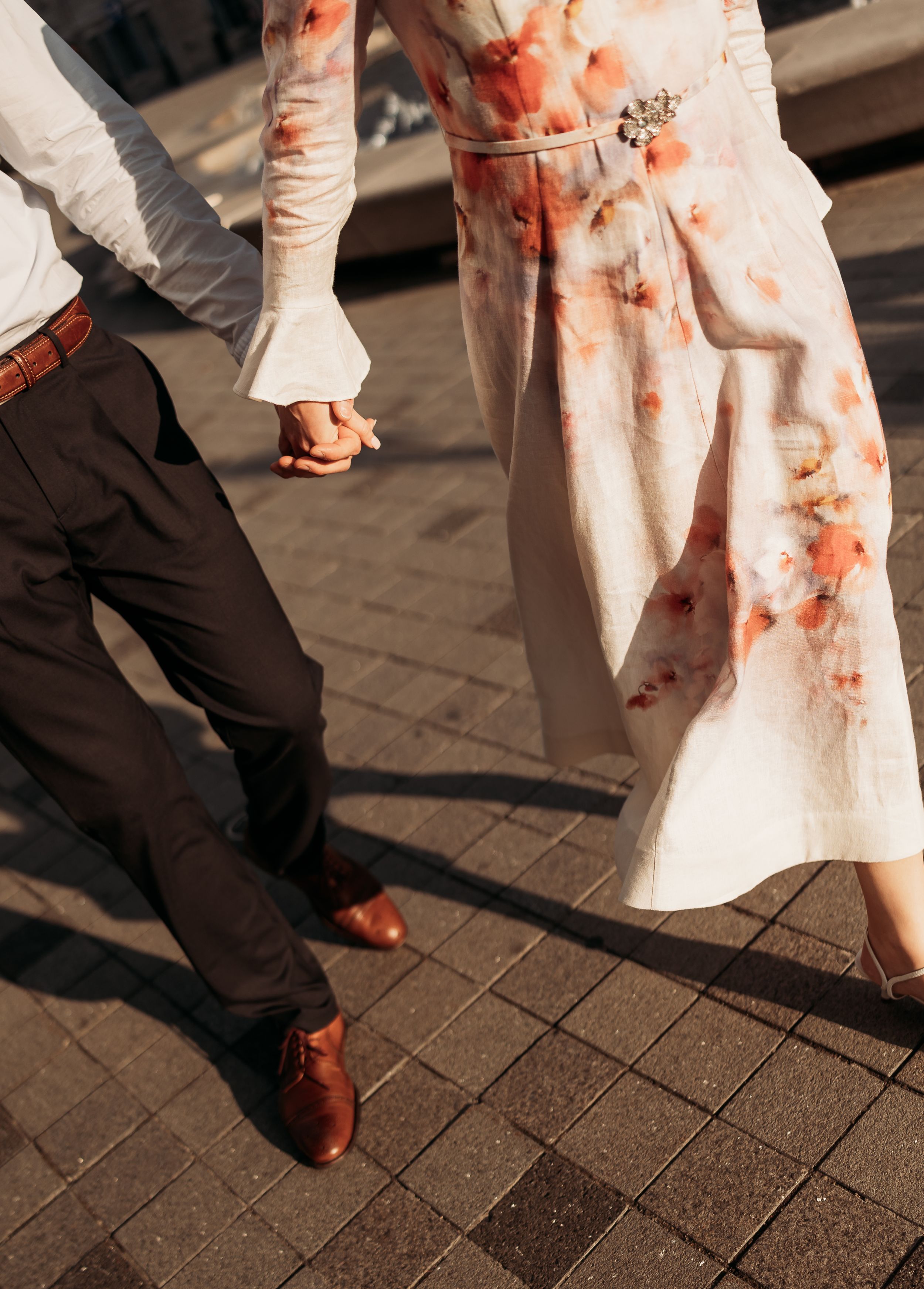 Couple holding hands walking on cobblestone, woman in floral dress, man in dark trousers and brown leather shoes.