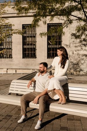 Couple relaxing on a white bench outdoors, woman in white dress sitting behind man in casual beige outfit, sunny urban setting.