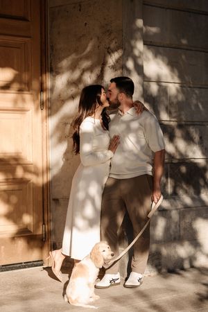 Couple sharing a kiss outdoors with a golden retriever puppy on a leash against a sunlit stone wall.