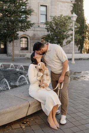 Couple sharing a kiss by a fountain, woman in white dress holding a golden puppy, urban setting with historic building.
