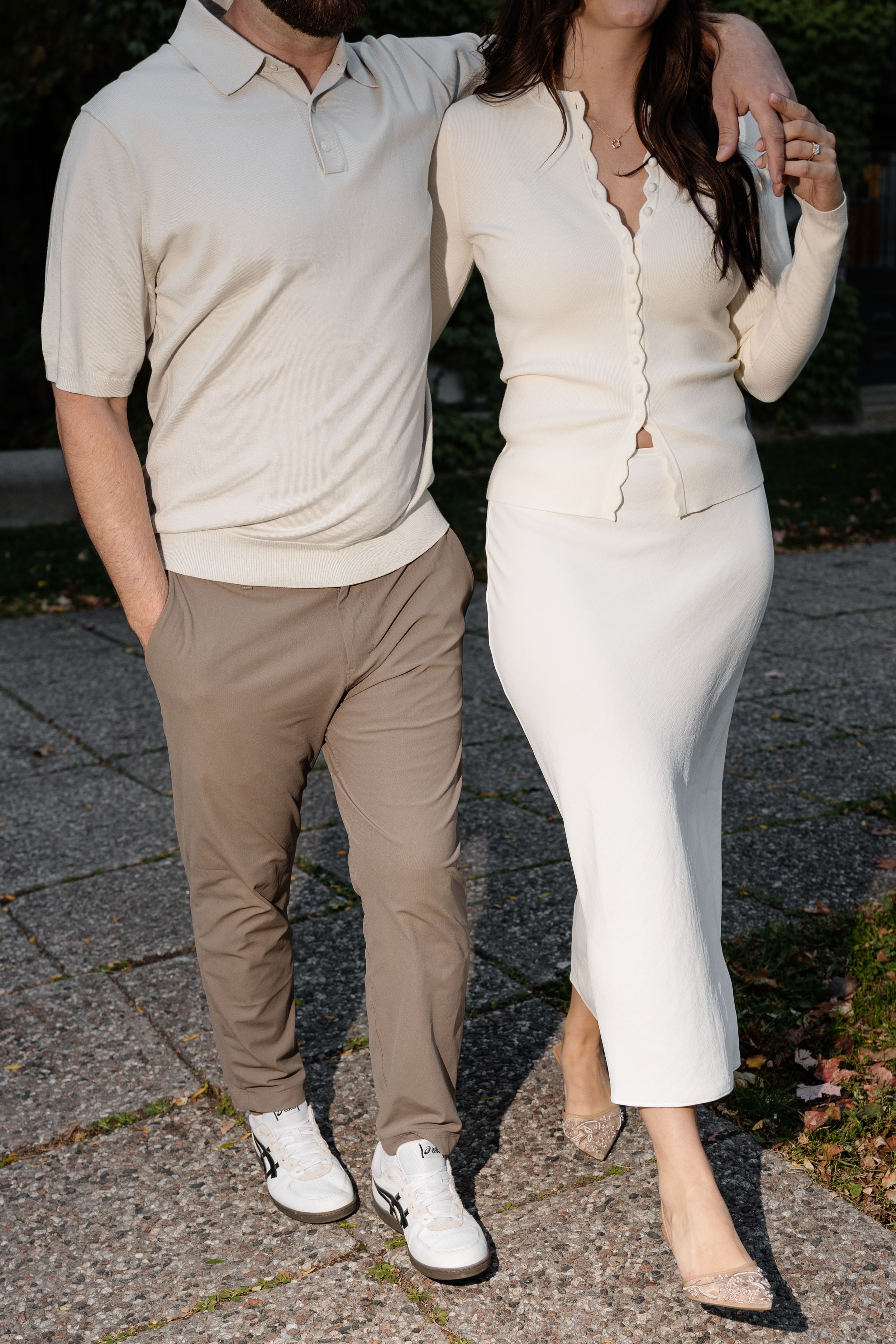Couple posing outdoors in coordinating neutral beige outfits, man in polo and chinos, woman in white knit skirt set.