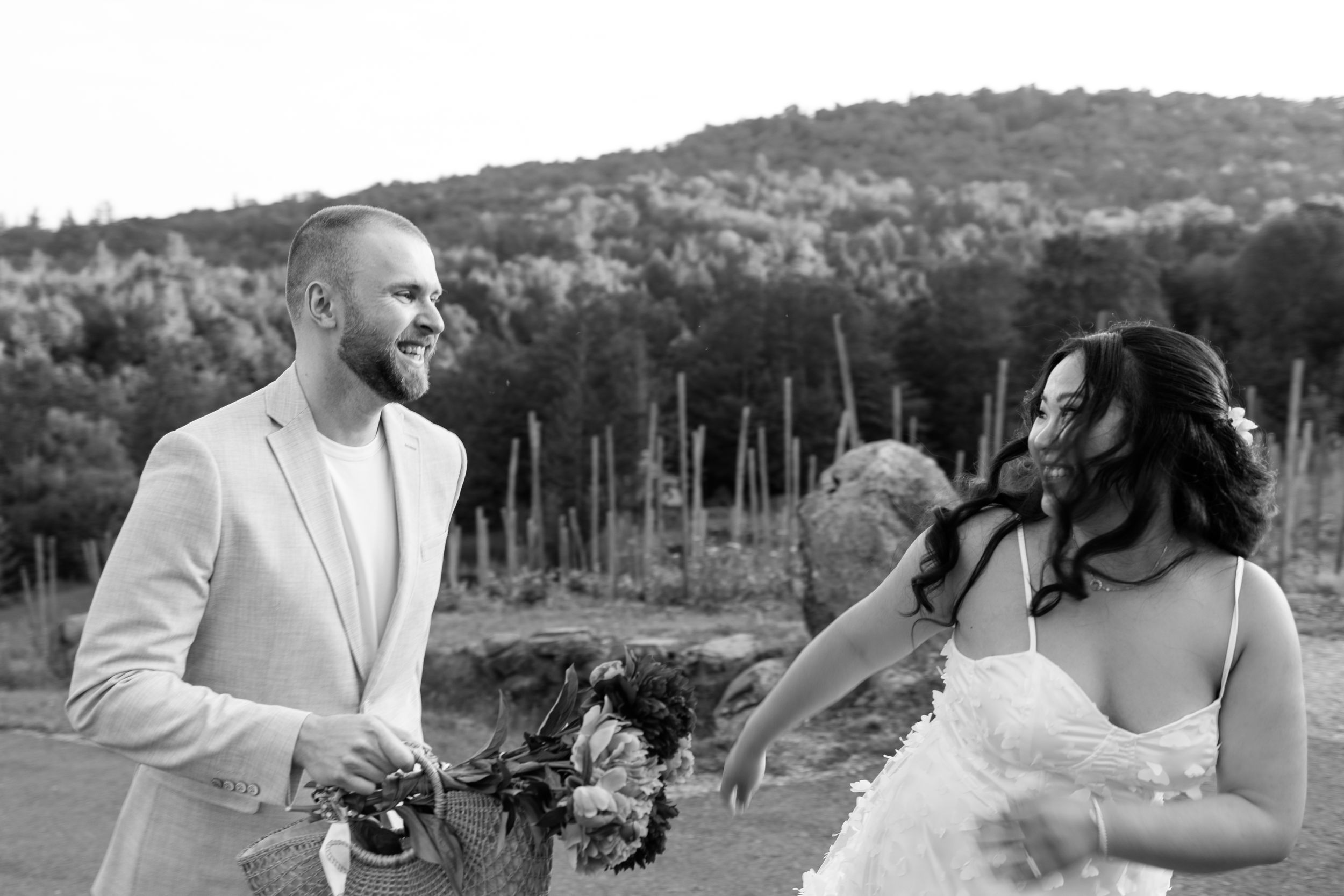 Black and white photo of a smiling groom in a light suit and bride in a floral dress outdoors at a vineyard wedding.