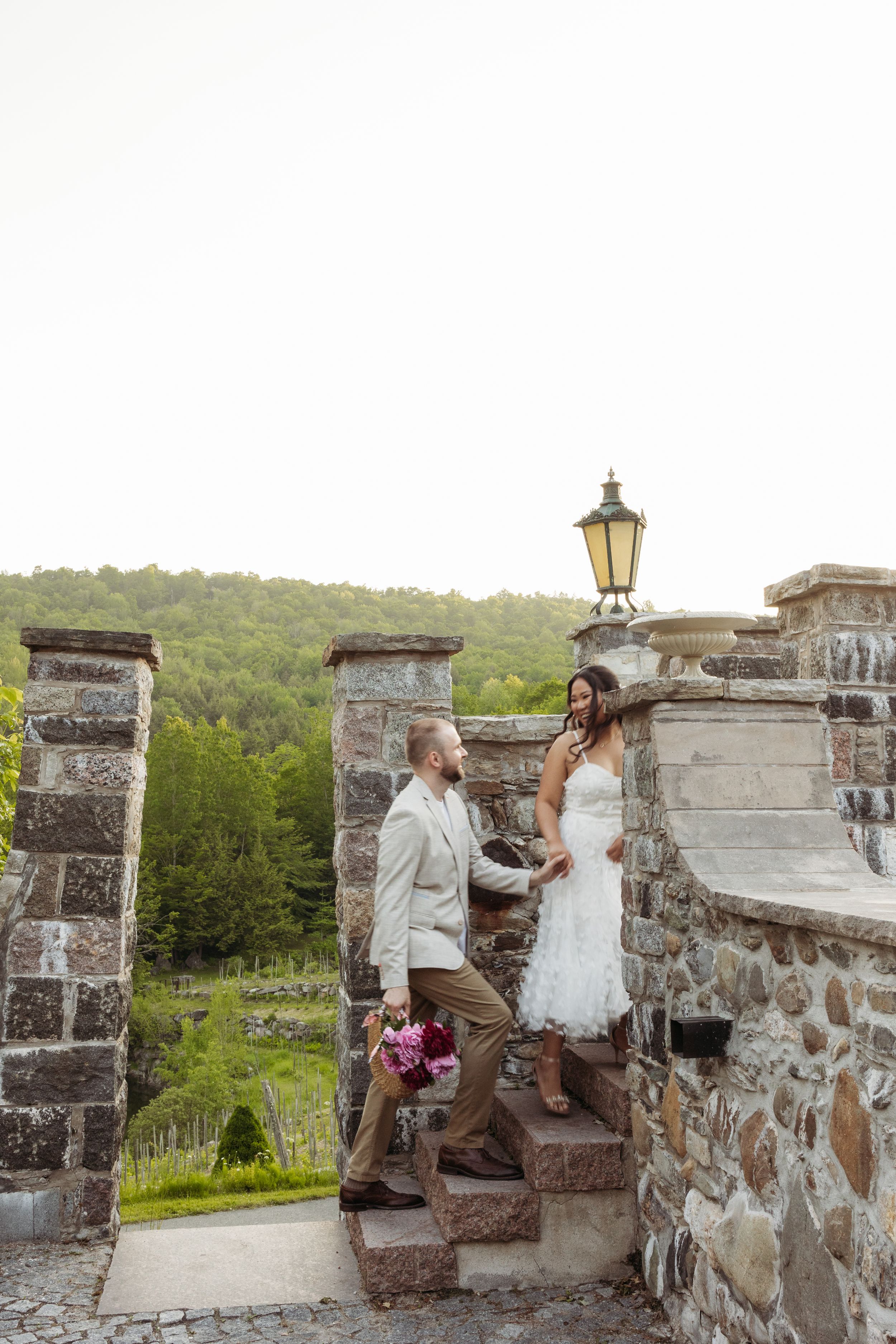 Bride and groom holding hands on stone steps at a rustic outdoor venue with lush green hills in the background.