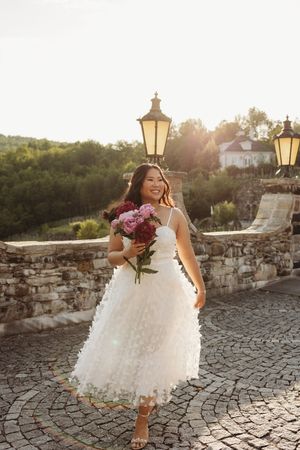 Smiling bride in white lace midi dress holding pink bouquet on cobblestone terrace at golden hour sunset.