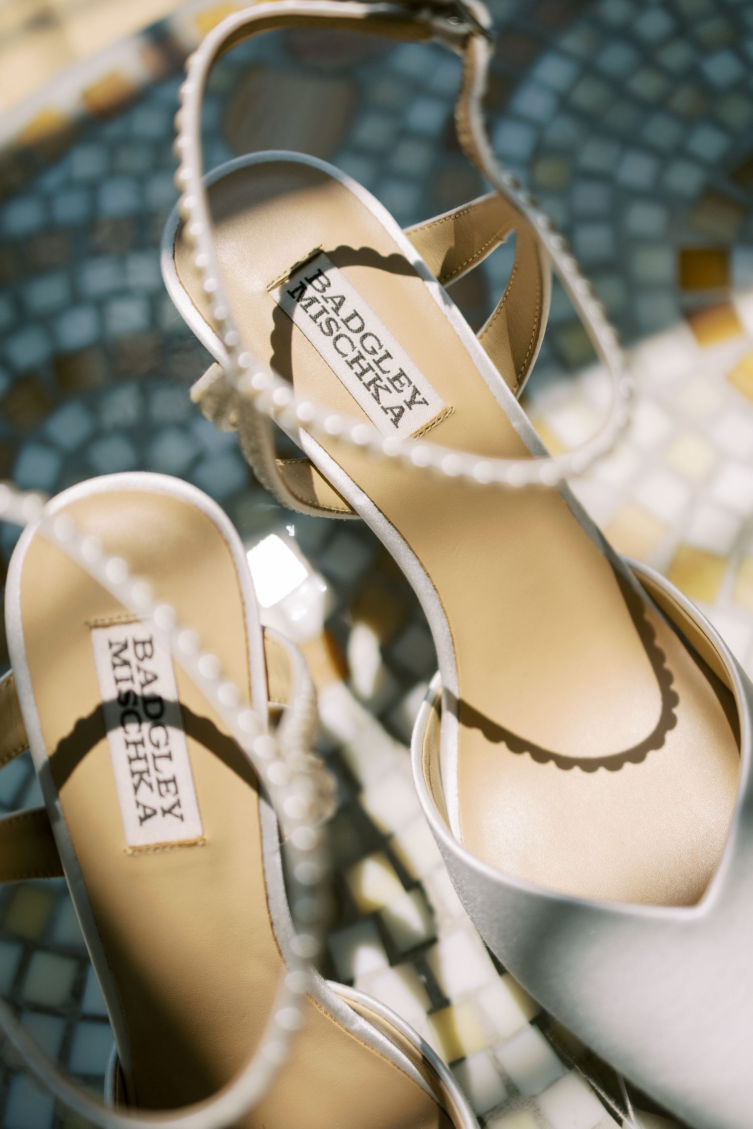 Badgley Mischka white satin bridal heels with pearl ankle straps displayed on mosaic tile floor.