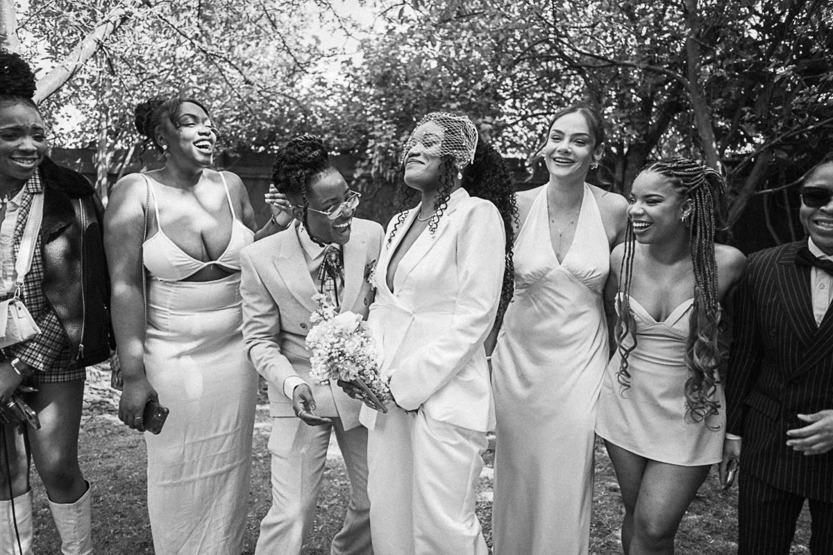 Black and white photo of a joyful wedding party in white outfits, laughing and celebrating outdoors.