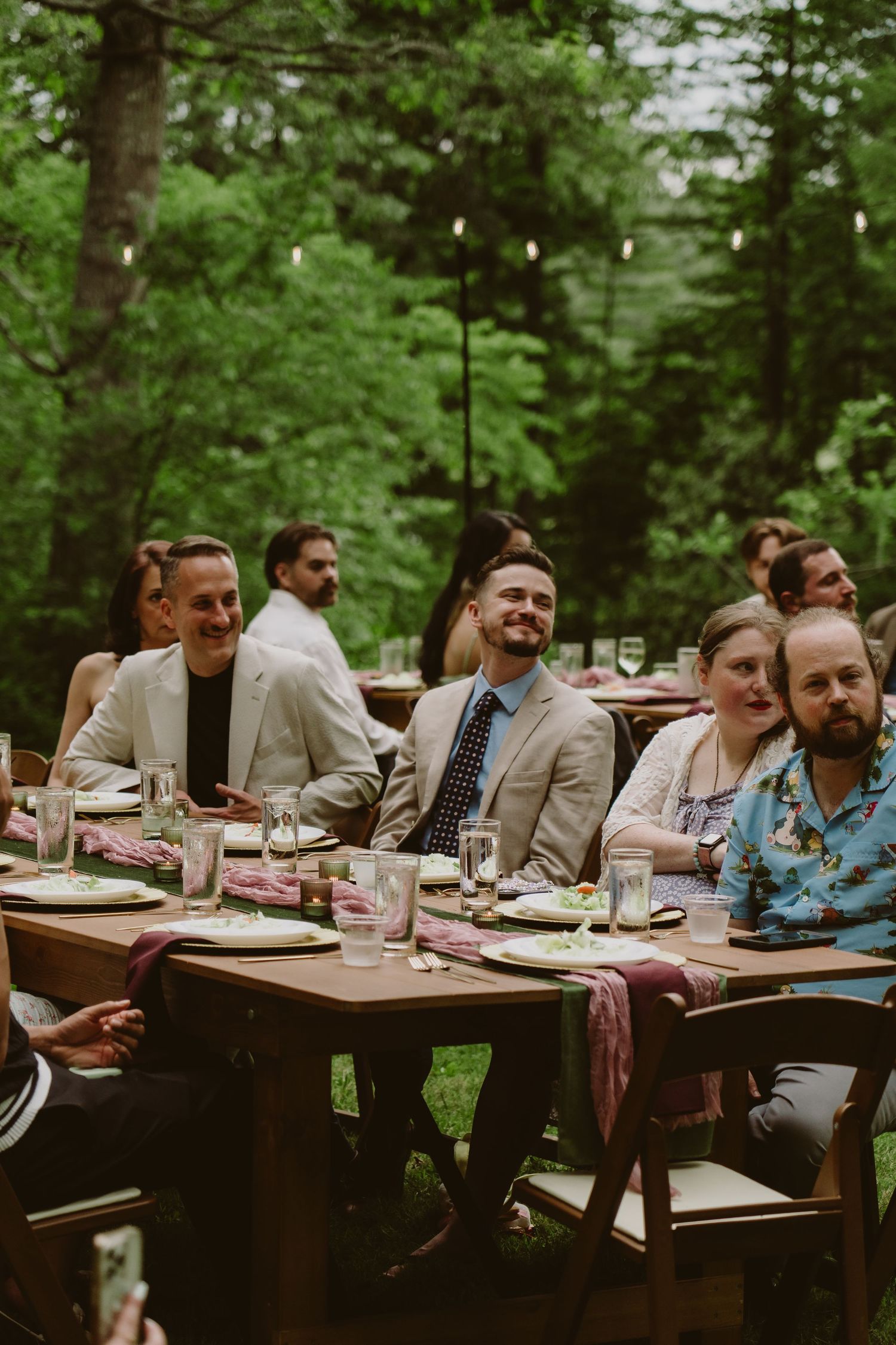 Rustic outdoor wedding reception table set in a sunlit forest with floral centerpieces, candles, and a sage green table runner.