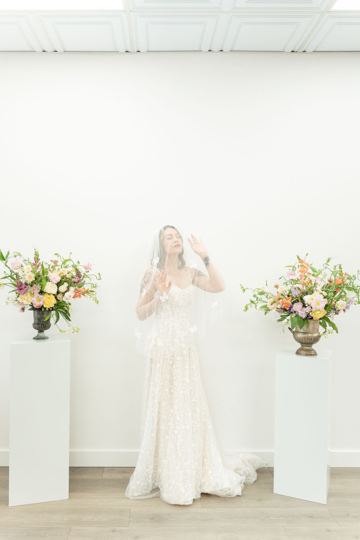 Bride in lace gown and veil poses between floral arrangements on pedestals in bright white studio.