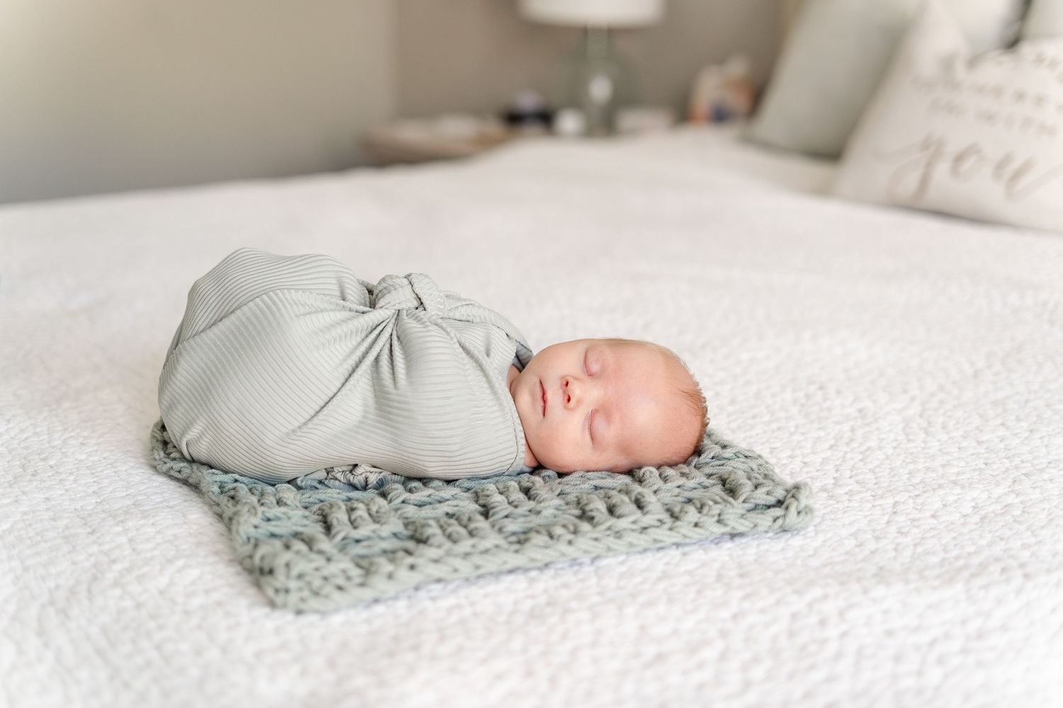 Newborn baby swaddled in sage green wrap sleeping peacefully on a knit blanket on a white bed.