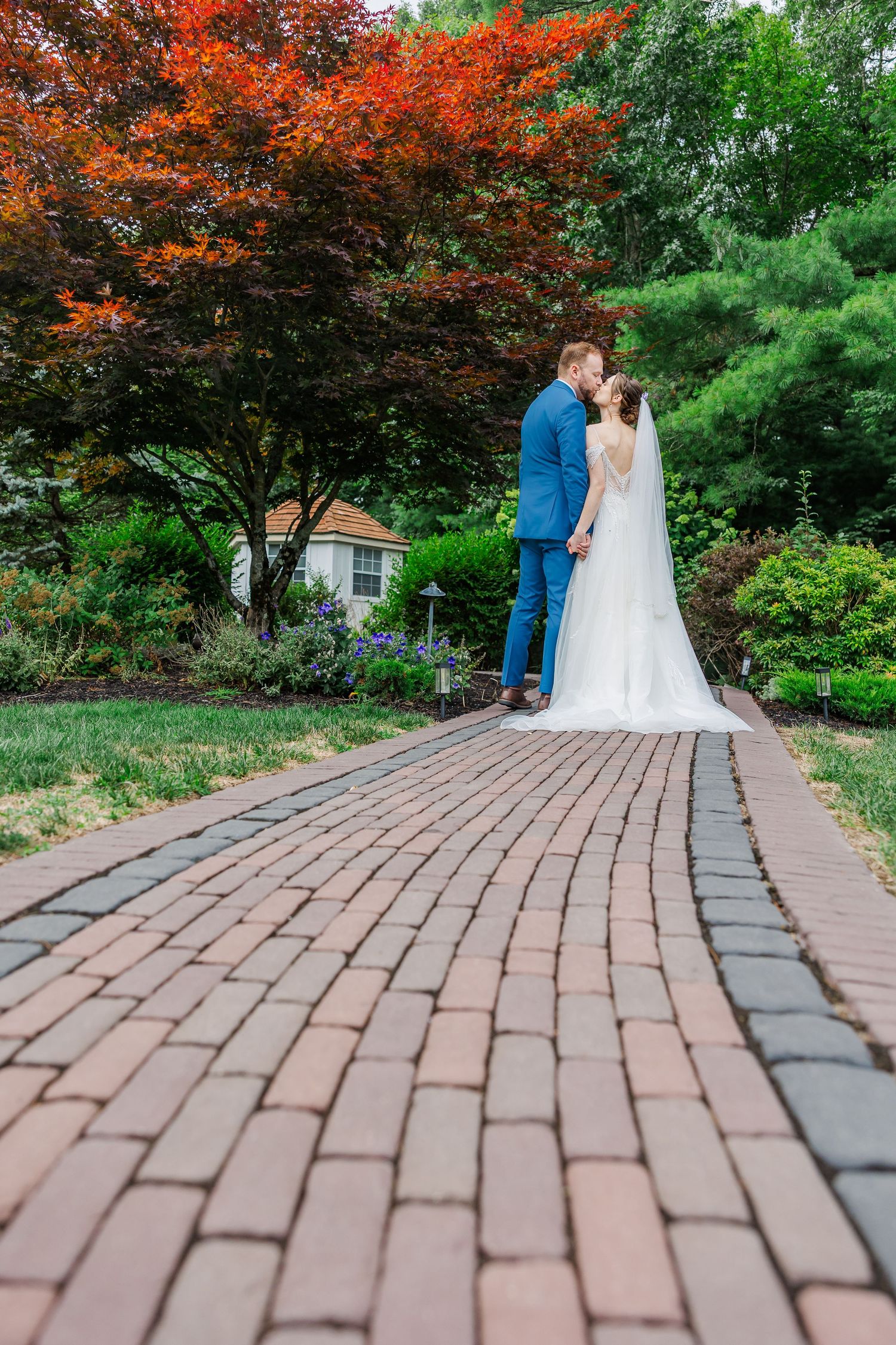 Bride and groom kissing on a brick pathway surrounded by lush gardens and vibrant red trees on their wedding day.
