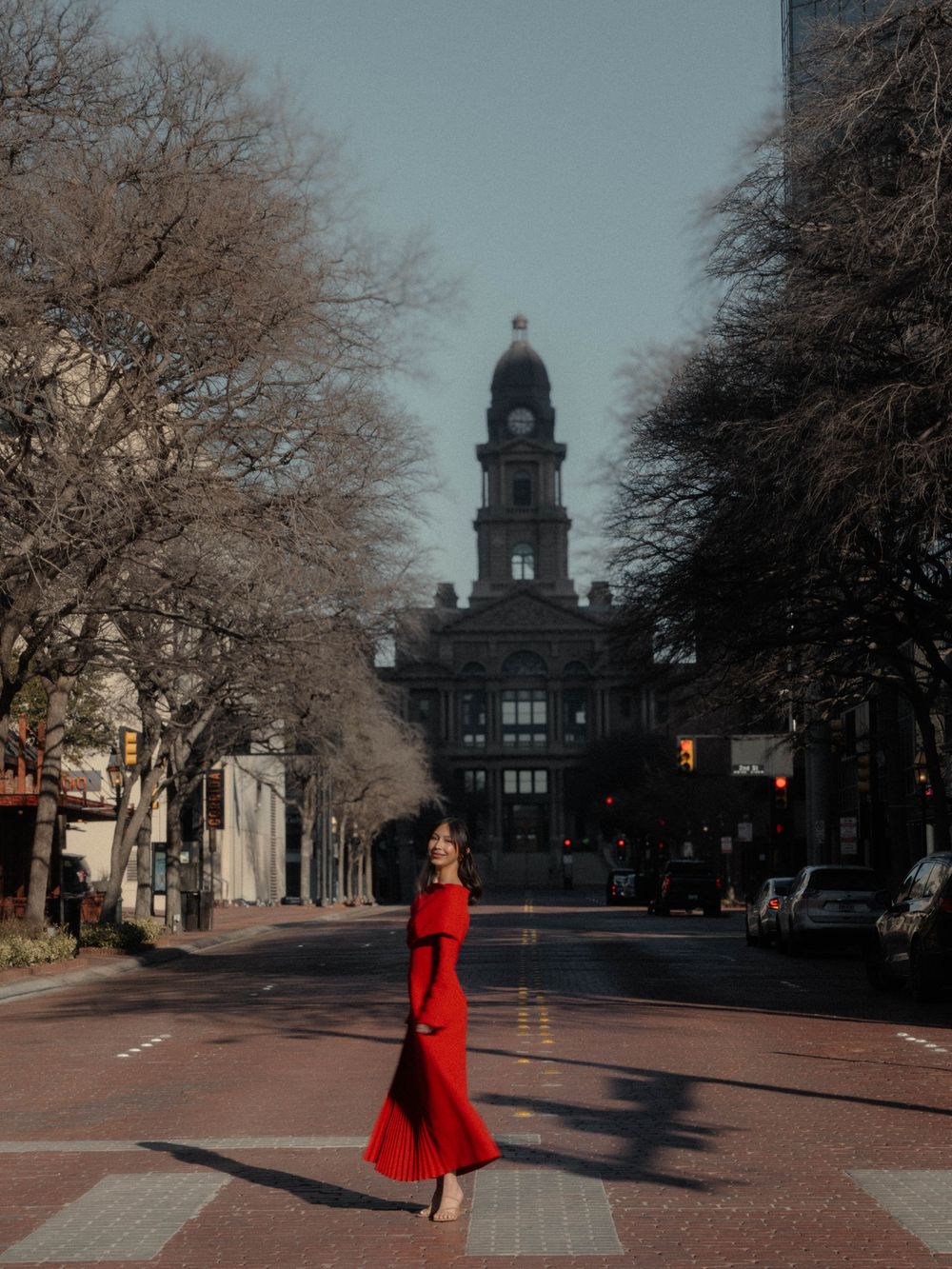 Woman in red off-shoulder maxi dress crossing a street with a historic clock tower behind her.