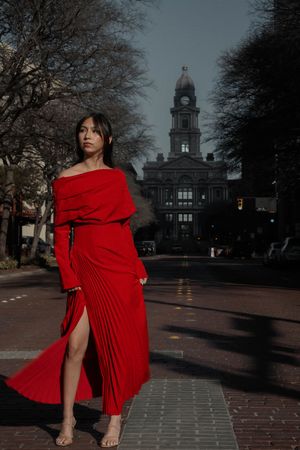 Woman in flowing red pleated dress standing on a street with a historic courthouse at dusk.
