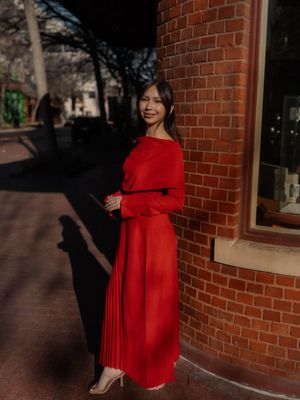 Smiling woman in red pleated maxi dress leaning against a sunlit brick building corner.