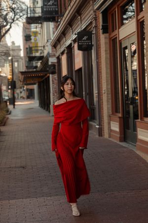 Woman in red off-shoulder pleated dress walking along a historic downtown brick sidewalk.