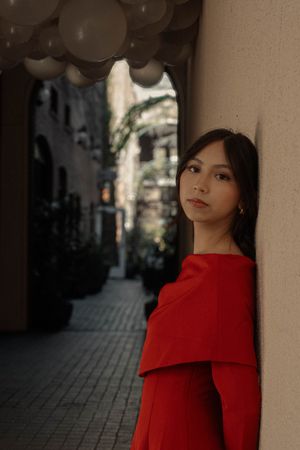 Young woman in red off-shoulder dress leaning against a wall under white balloon decorations.