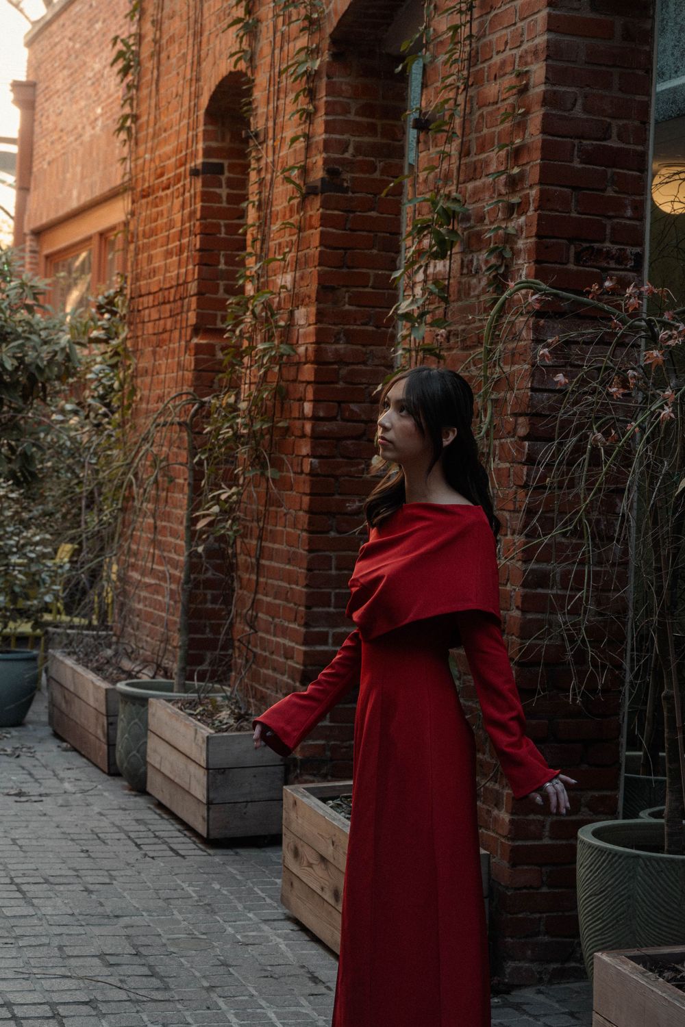 Woman in elegant red off-shoulder maxi dress posing in a brick courtyard with lush greenery.