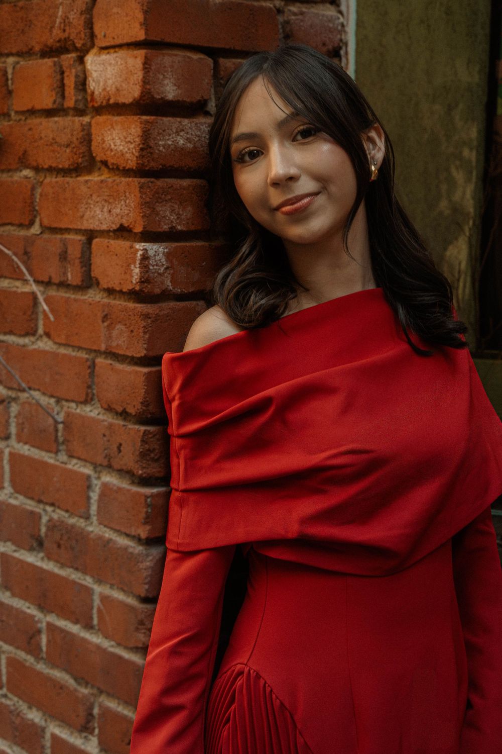 Young woman in red off-shoulder dress leans against rustic brick wall smiling softly.