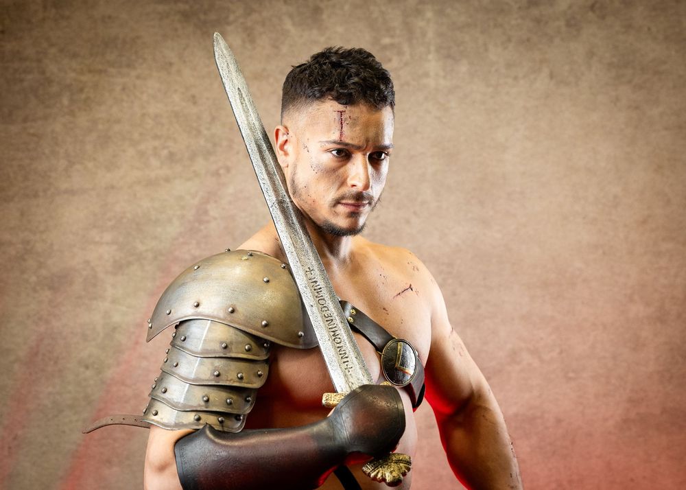 Muscular gladiator warrior poses with sword and armor against sepia background.