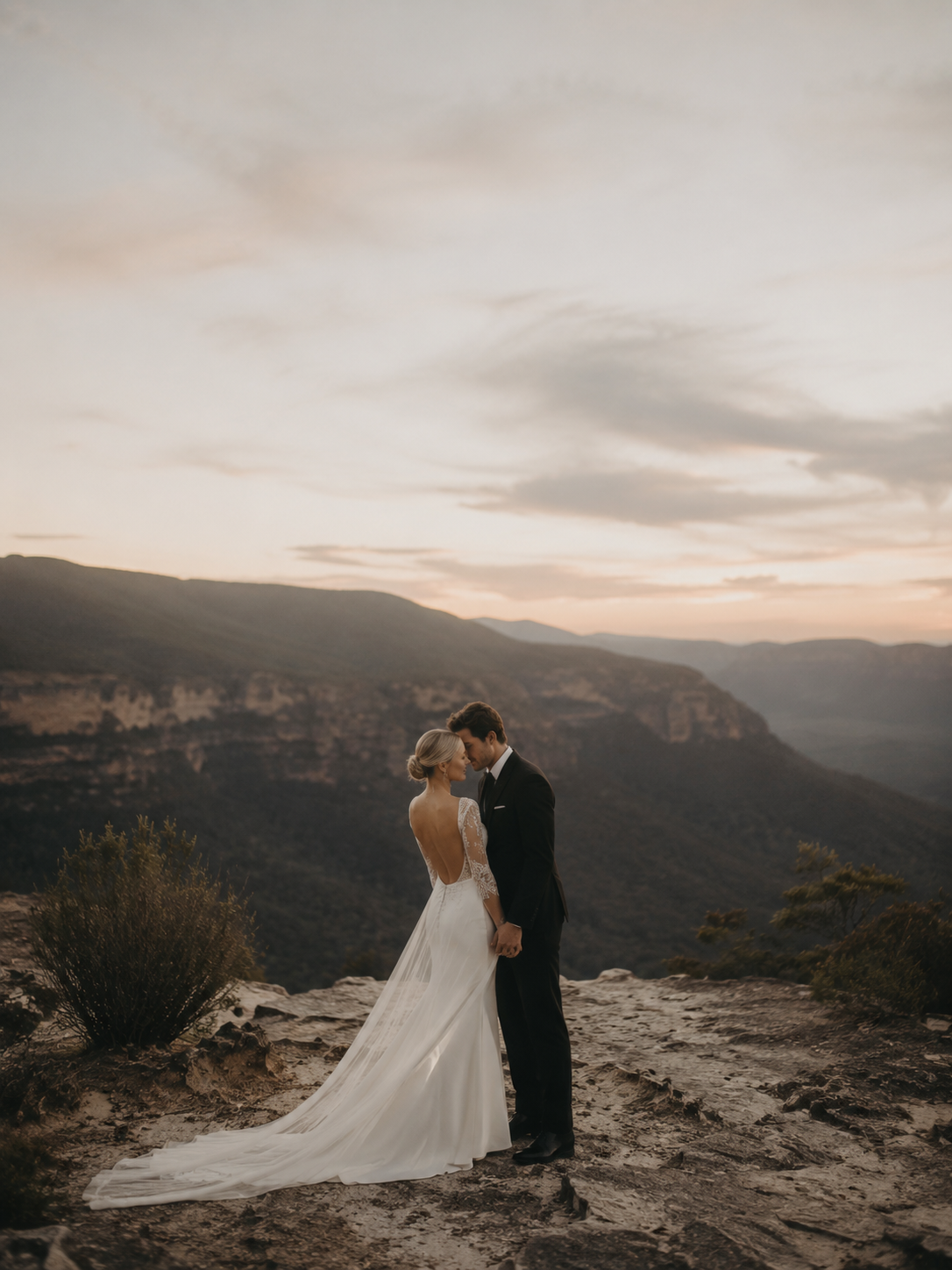 A bride and groom share an intimate moment on a rocky cliff overlooking a stunning mountain valley at sunset.