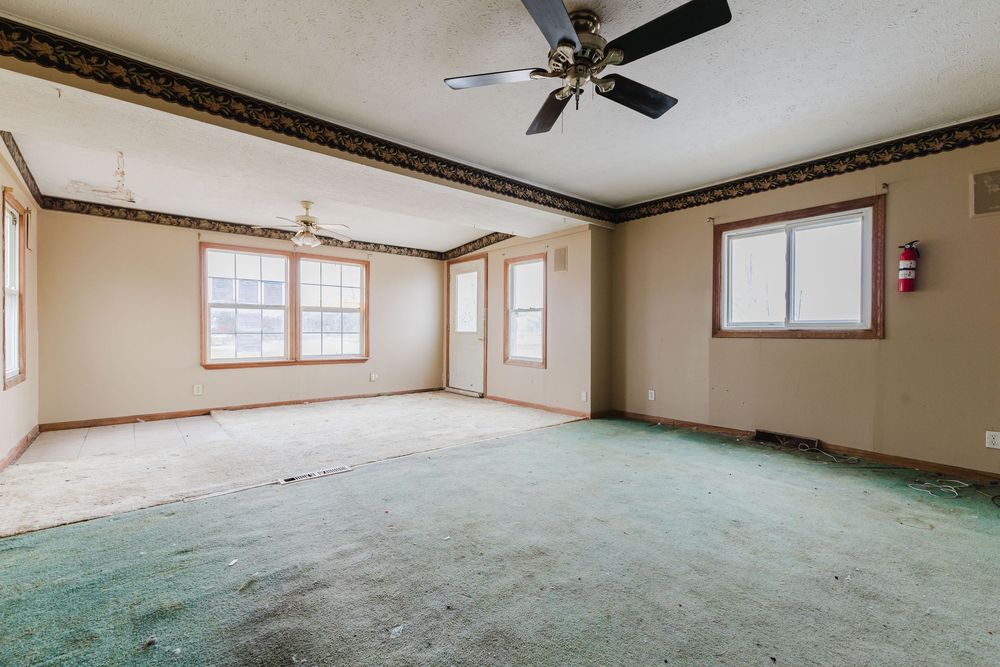 Spacious empty living room with worn green carpet, ceiling fan, and multiple windows.