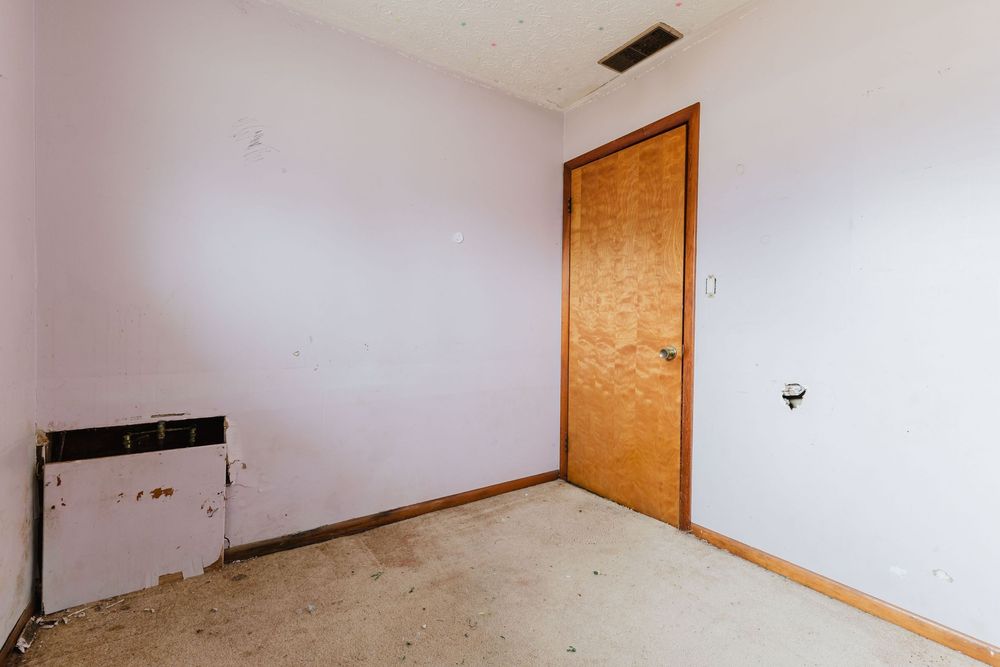 Empty room with bare concrete floor, wooden door, and white walls needing renovation.
