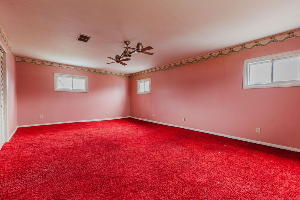 Spacious bedroom with bright red carpet, pink walls, ceiling fan, and two windows.