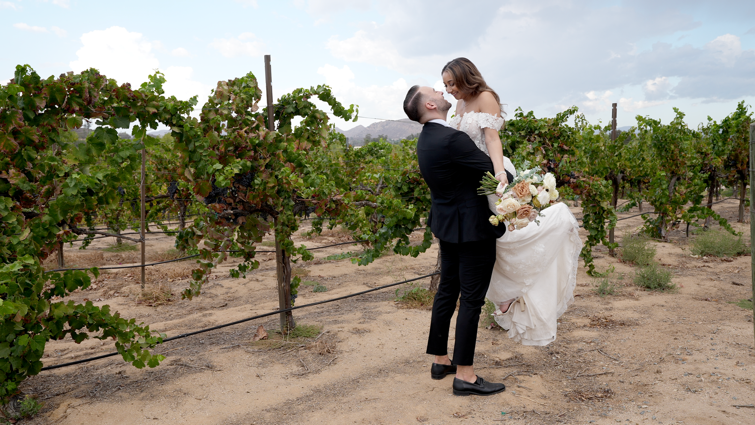 A romantic couple embraces while dancing in a lush vineyard on a cloudy afternoon.