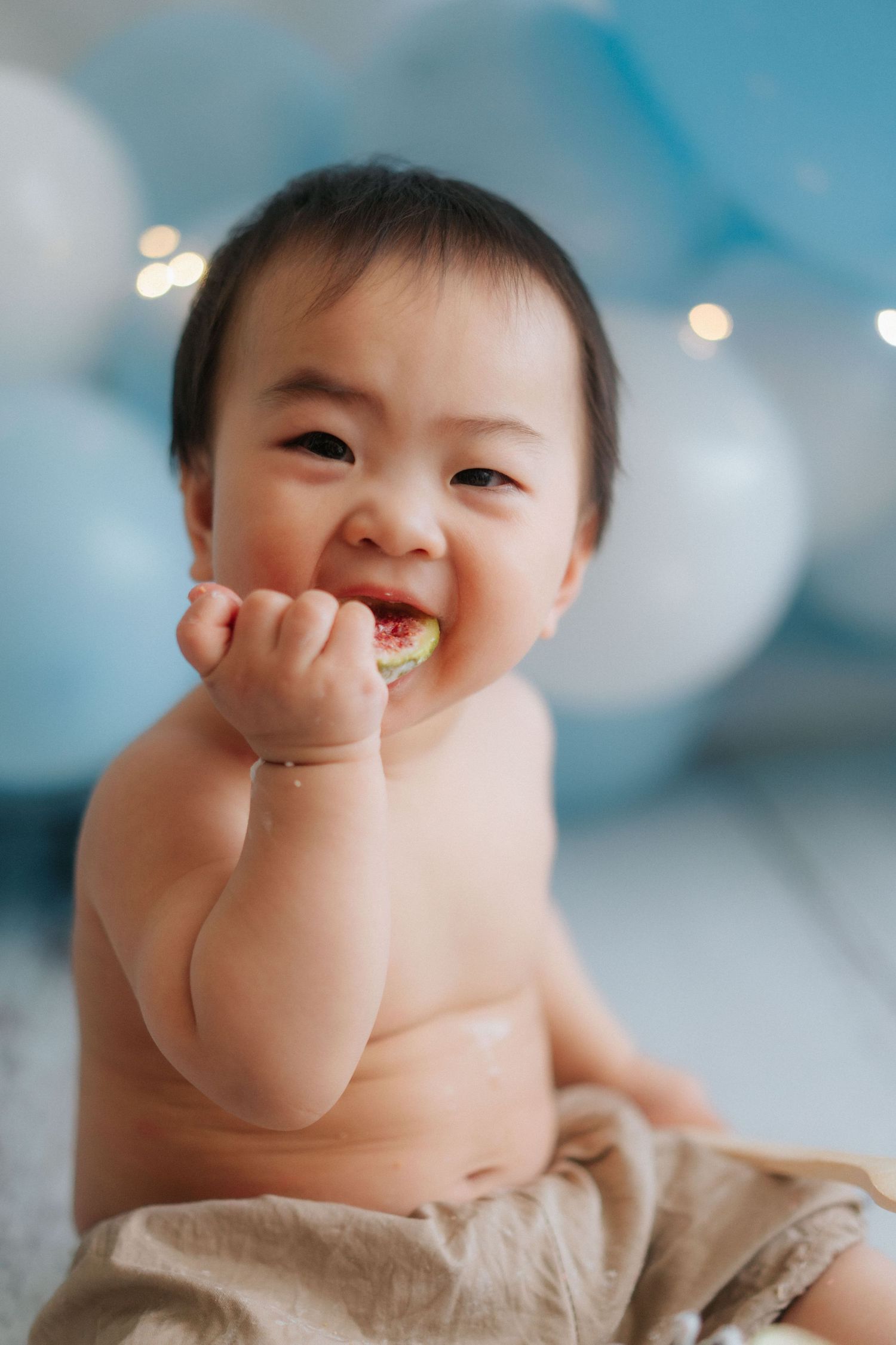 A baby laughs joyfully while covering their mouth with their hand against a soft blue background.