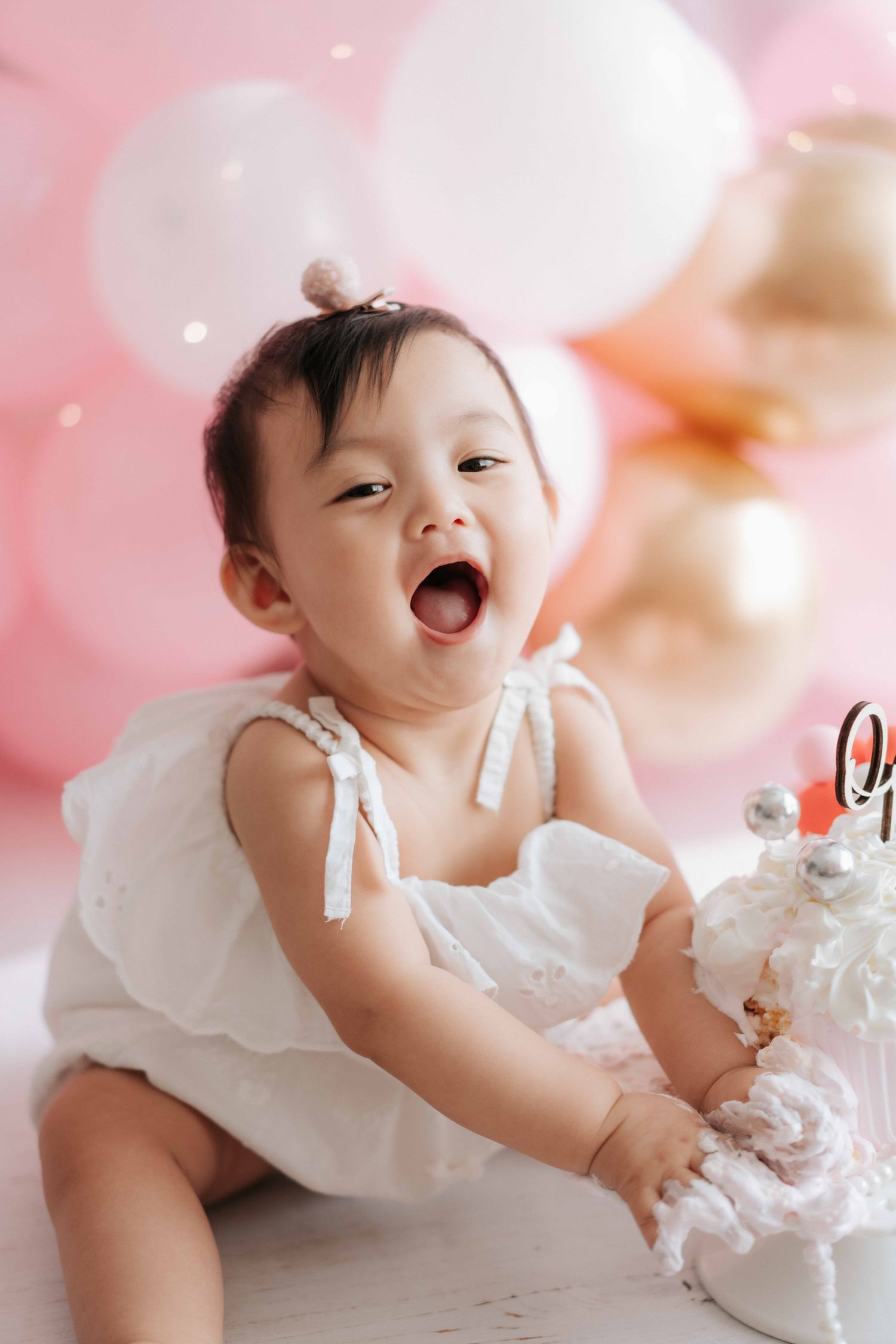 Baby in white dress laughs joyfully while sitting among pink and white balloons at a festive celebration.
