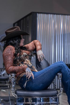 Female barber with tattoos wearing cowboy hat and floral top relaxing in salon chair holding scissors.