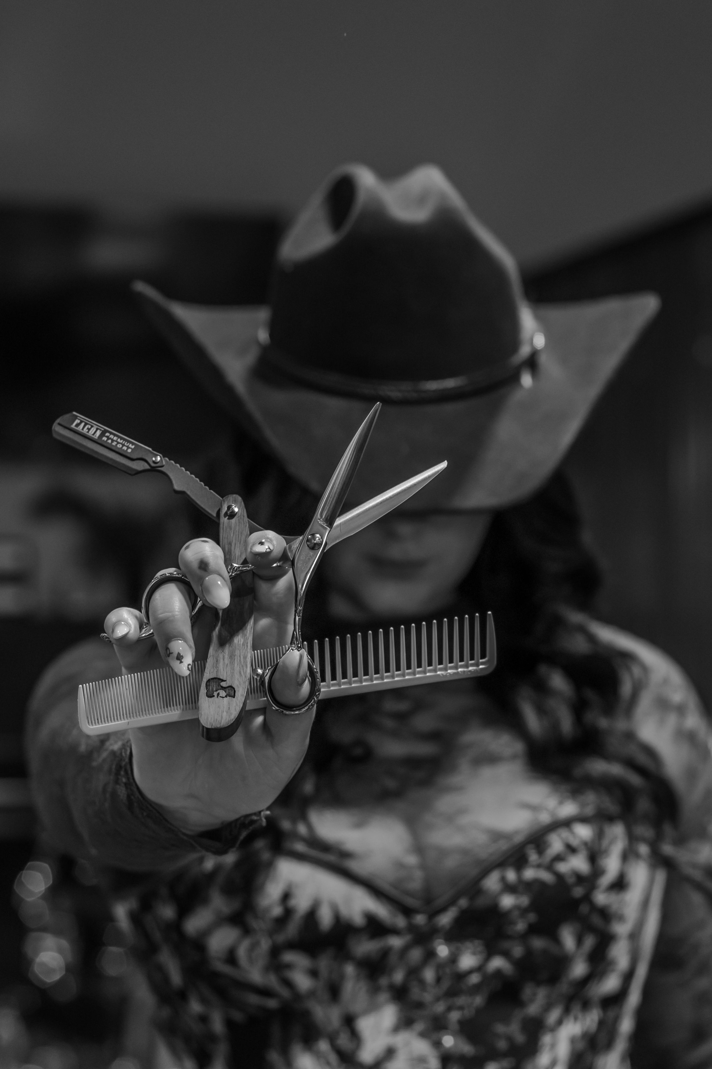 Black and white photo of a female barber in cowboy hat holding scissors, comb, and razor toward camera.