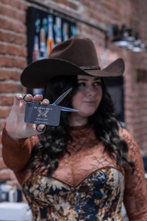 Female barber in cowboy hat holds business card and scissors toward camera in a brick barbershop.