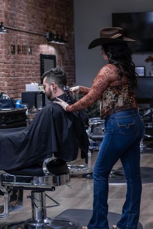 Female barber in cowboy hat cuts male clients hair in a stylish brick-walled barbershop interior.