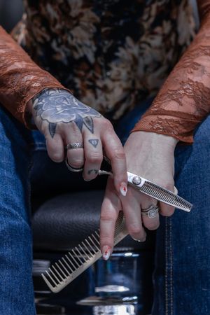 Close-up of tattooed female barber hands holding thinning shears and comb in a barbershop.