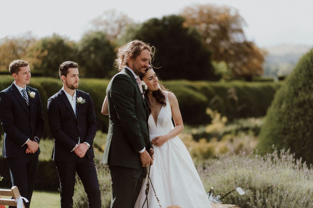 Wedding party gathers outdoors in a garden setting with manicured hedges and natural landscaping.