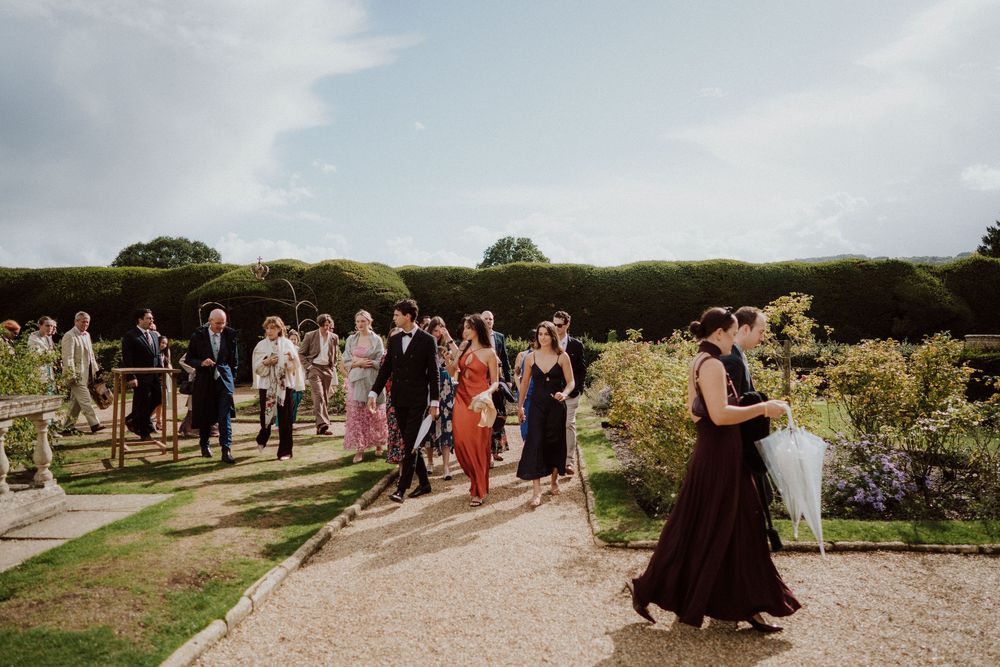 Wedding guests gather on a garden path surrounded by greenery and flowers.