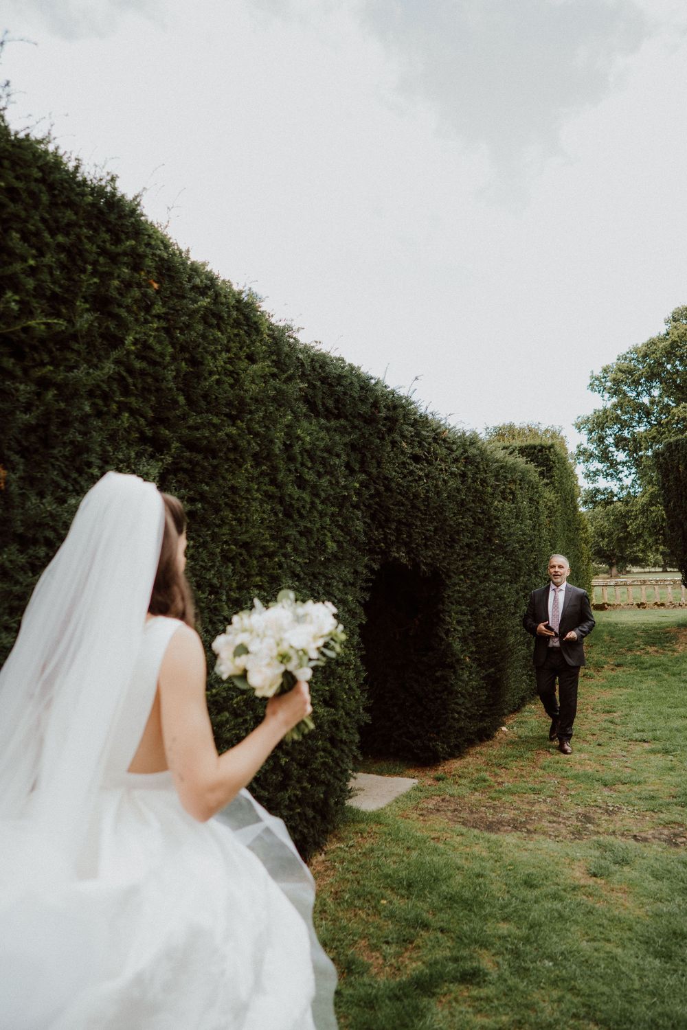 A bride in a flowing white dress and veil stands near tall hedges in an outdoor garden setting.