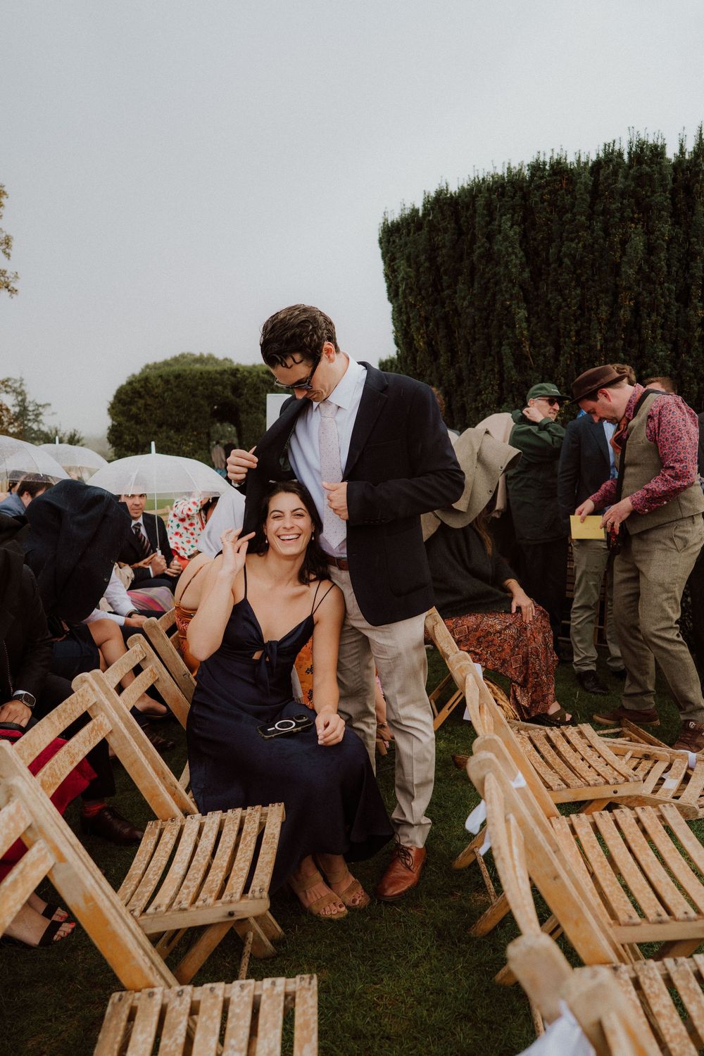 Wedding guests laugh while sitting on wooden folding chairs at an outdoor ceremony.
