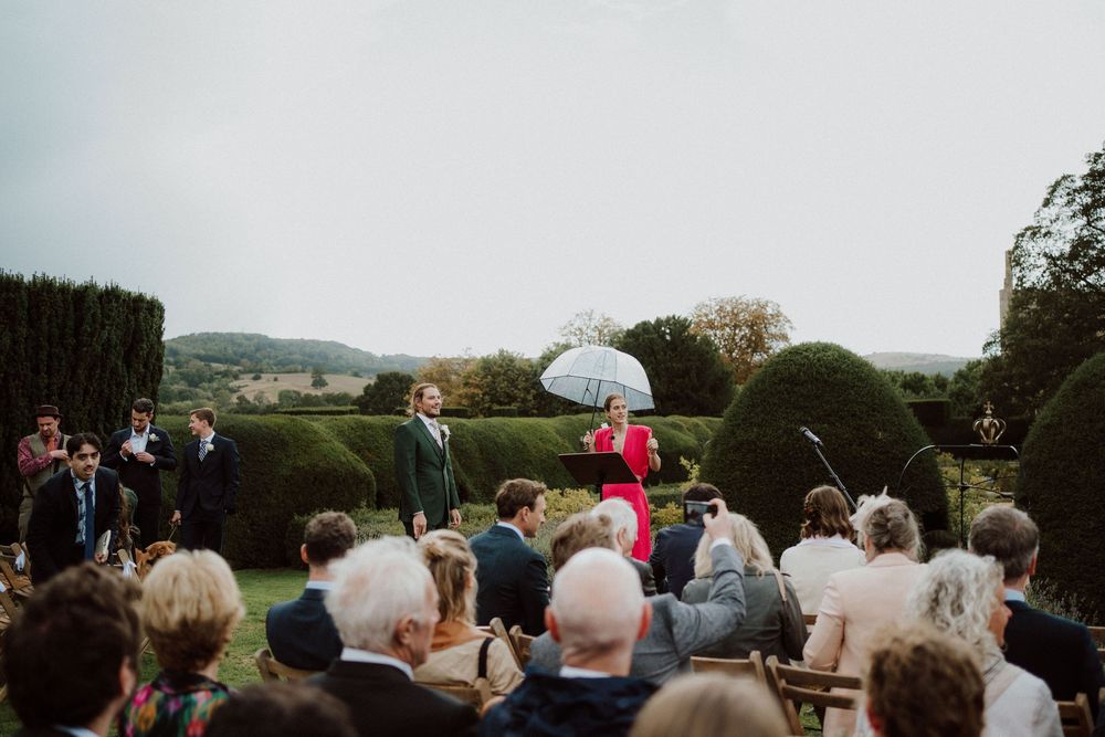 Panoramic view of wedding guests seated outdoors with umbrellas under cloudy skies.